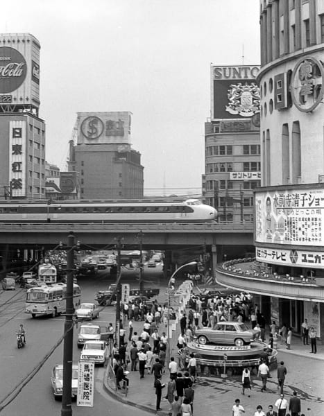 1964（昭和39）年に東京～新大阪駅間が開業した東海道新幹線。写真は67（昭和42）年、晴海通りと線路が交わる東京・有楽町を走り抜ける0系新幹線だ。街角には「東芝カラーテレビ」の広告やスカイライン・デラックスの車両展示も見られる